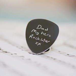 May include: A black guitar plectrum with the words "Dad My hero Rockstar xx" written in white. The plectrum is on a sheet of musical notation.