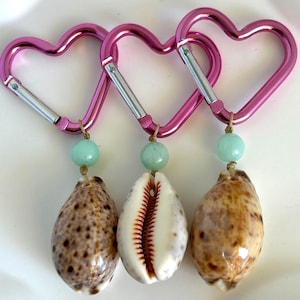 May include: Three pink heart-shaped carabiner clips with silver accents. Each clip has a light blue bead and a seashell pendant. The seashells are brown and white, with a variety of patterns and textures.