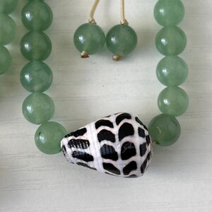 May include: A necklace featuring light green, round beads and a seashell pendant with a black and white pattern. The necklace also has two green bead accents and a beige string.