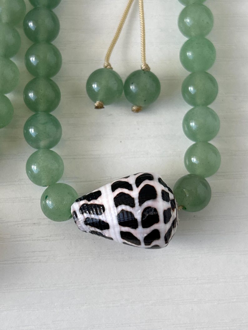 May include: A necklace featuring light green, round beads and a seashell pendant with a black and white pattern. The necklace also has two green bead accents and a beige string.