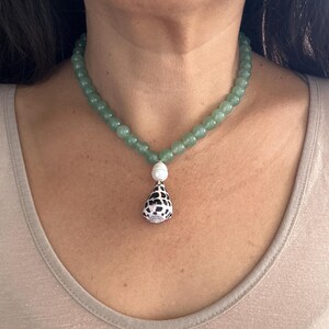 May include: A green beaded necklace with a pendant. The necklace features small, round, light green beads. The pendant is a white shell shape with black spots, and a white pearl bead. The necklace is worn around the neck.