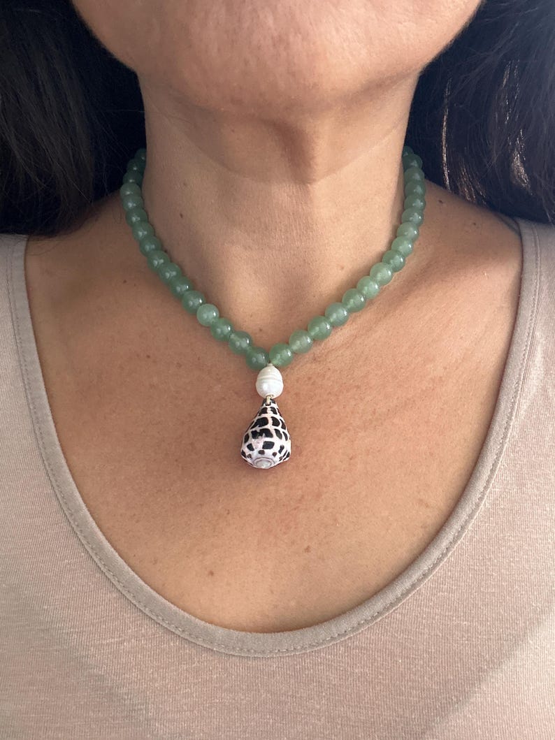 May include: A green beaded necklace with a pendant. The necklace features small, round, light green beads. The pendant is a white shell shape with black spots, and a white pearl bead. The necklace is worn around the neck.