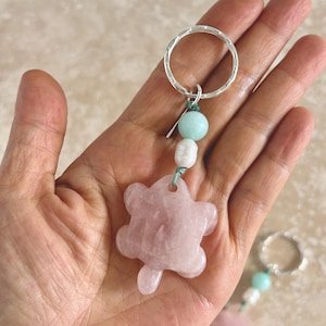 May include: A keychain featuring a carved light pink rose quartz turtle pendant suspended from a silver-toned metal ring. The turtle is attached by a green cord adorned with a smooth pale green round bead and a small white oval freshwater pearl.