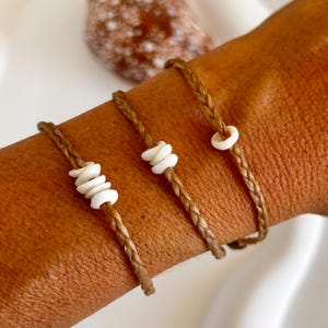 Puede incluir: Tres pulseras trenzadas marrones con cuentas de concha blanca. Las pulseras se llevan en la muñeca de una persona.
