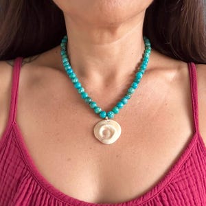 May include: A beaded necklace featuring round, turquoise-colored stones with dark veining, strung together to form a circular chain. A single, flat, off-white circular shell pendant with a spiral center design hangs from the center of the beads.