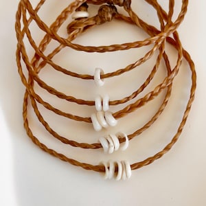 May include: A set of five brown braided bracelets with white shell beads. The bracelets are stacked on top of each other, with the beads increasing in size as they go down the stack.