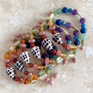 Puede incluir: Una colección de pulseras superpuestas con una mezcla de cuentas de piedras preciosas de colores en tonos azul, morado, verde, naranja y rosa, acentuadas con conchas cónicas con manchas negras y blancas sobre un cordón claro.