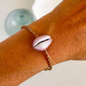 May include: A simple bracelet made with a brown braided cord and a pink seashell charm.