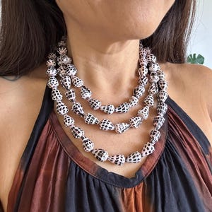 May include: A three-strand necklace featuring white and black spotted shell beads. The necklace is displayed against a backdrop of a brown and black tie-dye halter top. The beads are strung together to create a layered effect.