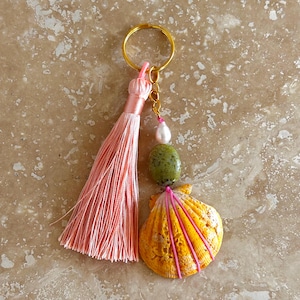 May include: A keychain featuring a gold-tone metal ring, a long light pink silk tassel, a small white pearl bead, a smooth olive green stone bead, and a yellow scallop shell wrapped with thin pink cord accents.
