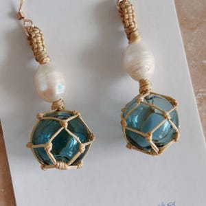 May include: A pair of handcrafted earrings featuring teal glass orbs encased in woven beige netting. Each earring includes a large, off-white pearl and gold-colored wire accents. The earrings dangle from gold-colored ear wires.