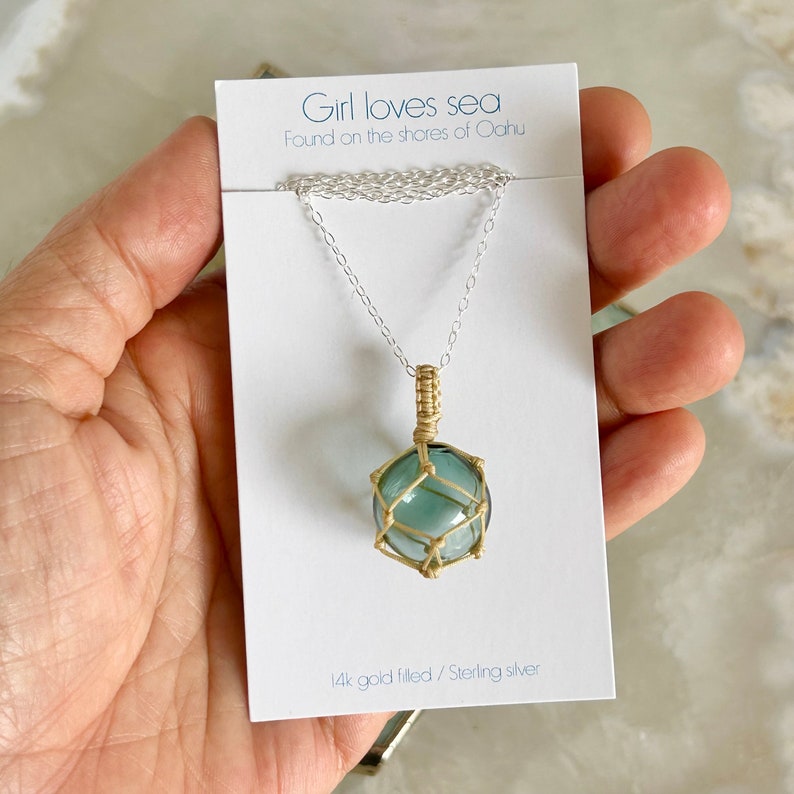 May include: A silver chain necklace with a gold-filled pendant. The pendant is a round, blue glass orb encased in a gold wire cage. The card reads "Girl loves sea Found on the shores of Oahu 14k gold filled/Sterling silver".