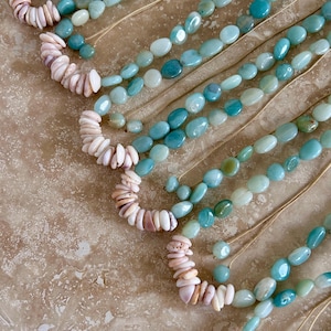 May include: Close-up of several strands of handmade jewelry. The necklaces feature a combination of light blue and turquoise oval-shaped beads, interspersed with sections of small, white and pink shell beads. The beads are strung on thin, natural-colored cords.