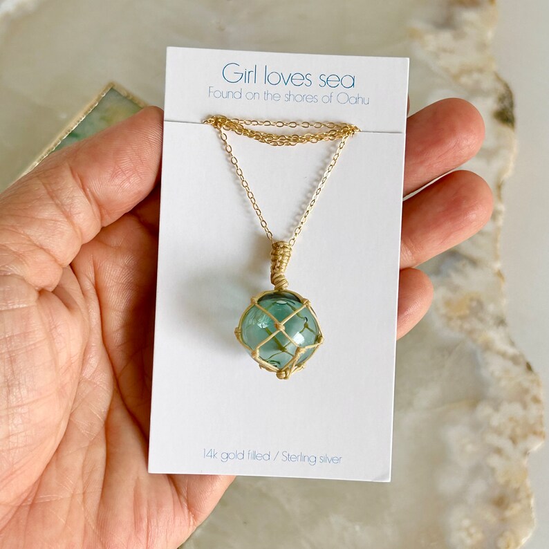 May include: A gold chain necklace with a green glass globe pendant encased in a woven wire cage. The card reads "Girl loves sea Found on the shores of Oahu 14k gold filled/Sterling silver".