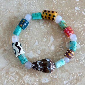 May include: A beaded bracelet featuring a variety of cylindrical and spherical beads in turquoise, yellow with black dots, red with white dots, white with black wavy lines, and a large brown cone-shaped shell bead interspersed with small translucent beads.