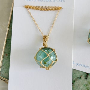May include: A gold chain necklace with a glass float pendant. The float is encased in a woven net and has a light blue color.