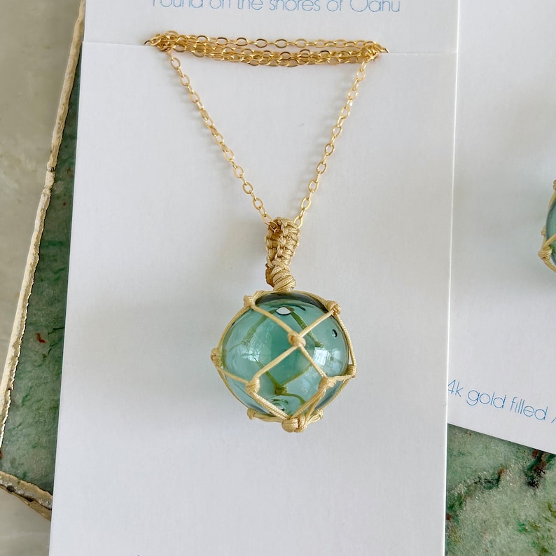 May include: A gold chain necklace with a glass float pendant. The float is encased in a woven net and has a light blue color.