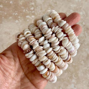May include: Several bracelets made of small, flat, white and tan seashells. The bracelets are stacked together, showing the natural variations in color and texture of the shells. The shells are strung together, creating a unique accessory.