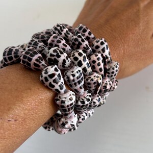 Peut inclure: Plusieurs bracelets faits de coquillages naturels. Les coquillages sont blancs avec des taches noires et une teinte ros&eacute;e. Les bracelets sont port&eacute;s sur un poignet.