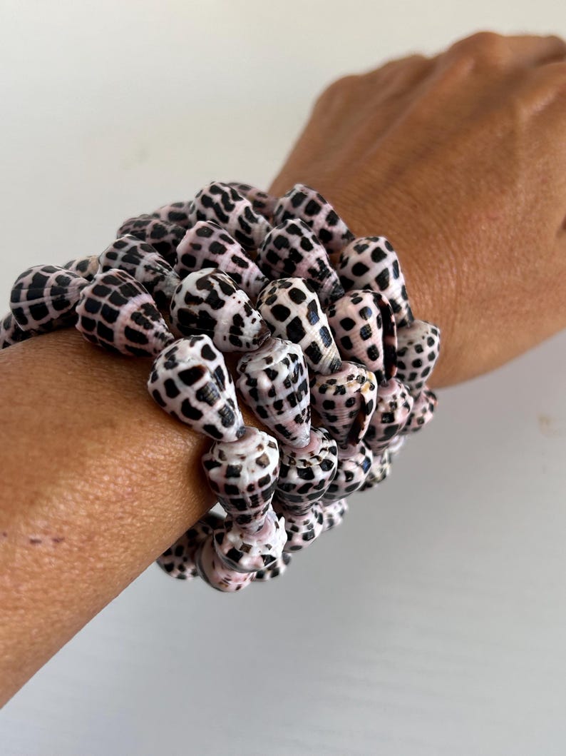 Peut inclure: Plusieurs bracelets faits de coquillages naturels. Les coquillages sont blancs avec des taches noires et une teinte ros&eacute;e. Les bracelets sont port&eacute;s sur un poignet.