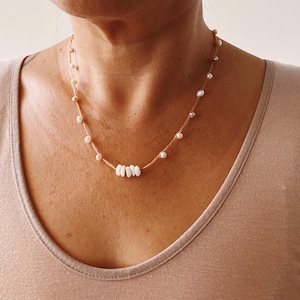 Puede incluir: Un delicado collar con pequeñas perlas redondas y cuentas blancas oblongas ensartadas en un fino cordón beige. El collar se muestra sobre un fondo neutro, resaltando la sencilla elegancia de la joya. Un accesorio perfecto para el uso diario.