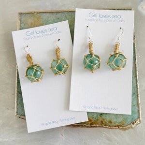 May include: A pair of earrings featuring a green glass ball encased in a woven straw net. The earrings are on a white card with the text "Girl loves sea Found on the shores of Oahu 14k gold filled/Sterling silver".
