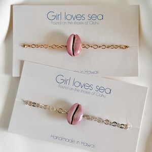 May include: Two bracelets with pink seashells and gold and silver chains. The bracelets are displayed on white cards with the text "Girl loves sea" and "Found on the shores of Oahu".
