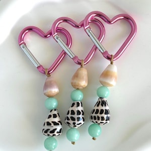 Puede incluir: Tres mosquetones rosas en forma de corazón con detalles plateados. Cada mosquetón tiene una cadena de cuentas y conchas. Las cadenas incluyen una concha rosa claro, una cuenta verde claro y una concha con estampado en blanco y negro.
