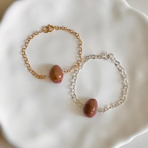 May include: Two bracelets with oval-shaped, reddish-brown beads. One bracelet has a gold-colored chain, and the other has a silver-colored chain. Both bracelets are on a white, scalloped plate.