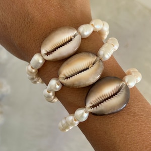 Op de afbeelding: Drie armbanden met witte parel kralen en kauri schelpen. De schelpen zijn bruin en hebben een natuurlijke, organische textuur.