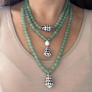 May include: Three-strand necklace featuring light green beads. Each strand has a different length. The necklace is adorned with black and white shell pendants and a white pearl. The jewelry is worn on a person.