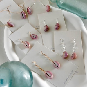 May include: A set of pink cowrie shell earrings on display cards. The earrings are available in gold filled, sterling silver, and 14k gold.