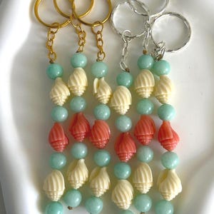 May include: Keychains with gold and silver rings, featuring strands of turquoise beads, cream-colored shell beads, and coral-colored shell beads. The keychains are arranged on a white surface.