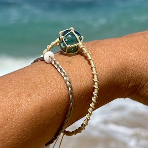 Op de afbeelding: Twee armbanden om een pols. Een armband is gemaakt van gevlochten beige koord. De andere armband is gemaakt van beige koord met een teal glazen kraal.