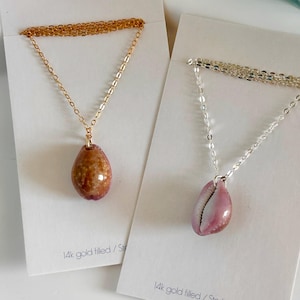 May include: Two necklaces with shell pendants. The necklace on the left has a gold chain and a brown shell pendant. The necklace on the right has a silver chain and a pink shell pendant. Both necklaces have the text "14k gold filled / Sterling Silver" on the card.