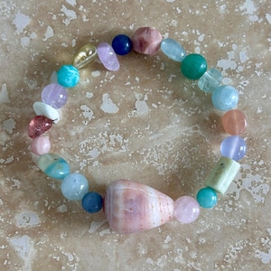 May include: A stretch bracelet featuring a variety of multicolored gemstone beads in shades of pink, blue, green, yellow, and purple. The bracelet includes round, faceted, and irregular shapes, with a large, central, cone-shaped pink seashell bead.