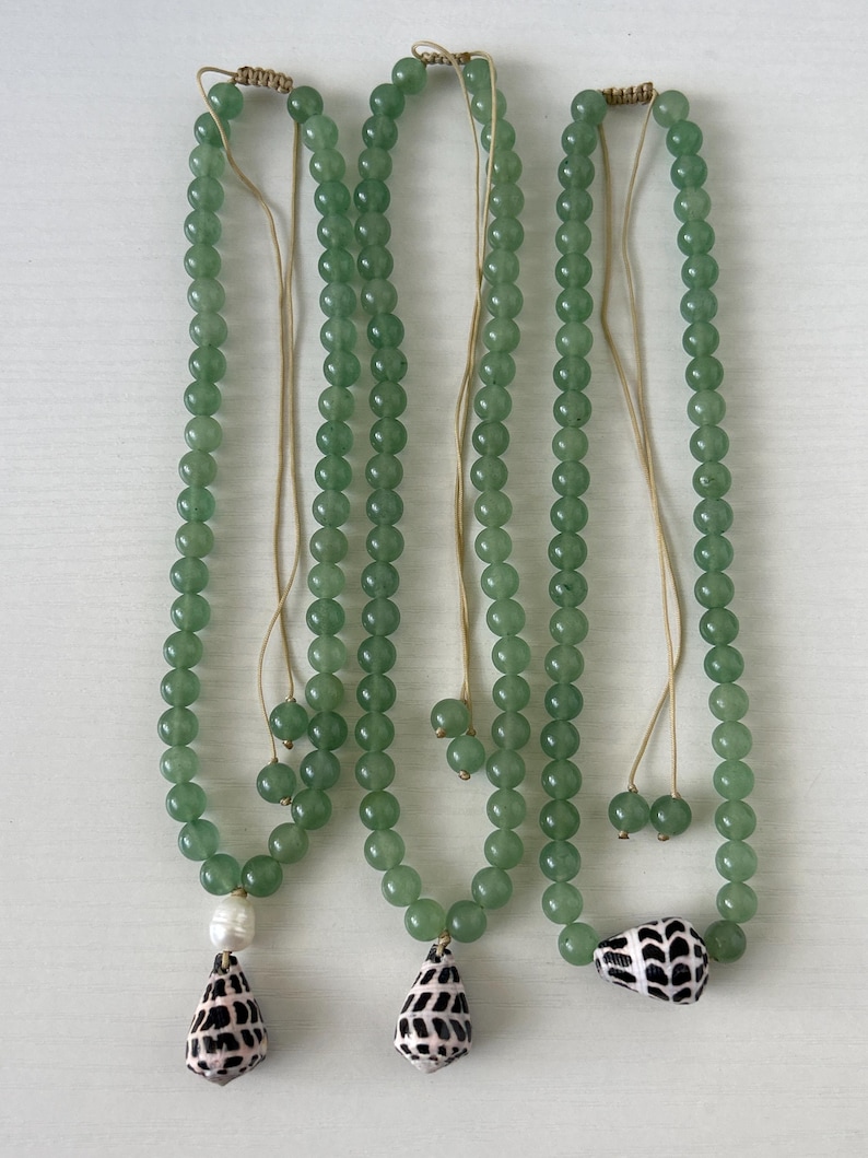 May include: Three green beaded necklaces with seashell pendants. The necklaces have a light green hue and are strung with small, round beads. Each necklace features a different seashell pendant, with one featuring a white pearl.