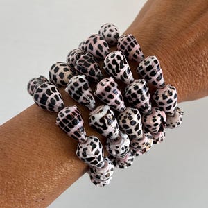 Può includere: Una pila di braccialetti fatti di conchiglie naturali. I gusci sono bianchi con macchie nere e accenti rosa. I braccialetti sono indossati su un polso.