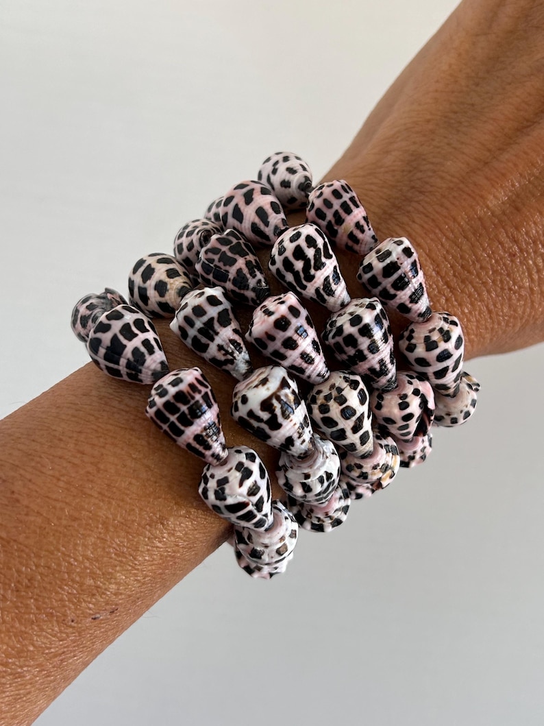 Peut inclure: Plusieurs bracelets faits de coquillages naturels. Les coquillages sont blancs avec des taches noires et des accents roses. Les bracelets sont port&eacute;s sur un poignet.