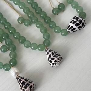May include: Close-up of several necklaces featuring light green beads. Each necklace has a unique pendant, including a seashell with a black and white pattern and a white pearl. The necklaces are displayed on a white surface.
