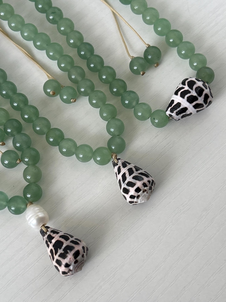 May include: Close-up of several necklaces featuring light green beads. Each necklace has a unique pendant, including a seashell with a black and white pattern and a white pearl. The necklaces are displayed on a white surface.