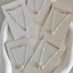 Könnte beinhalten: Fünf silberne Halsketten mit weißen Keramikringen auf weißem Hintergrund. Die Halsketten werden auf Karten mit dem Text "Sterling Silver" präsentiert.