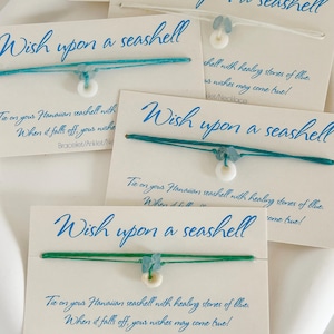 Puede incluir: Tres tarjetas con el texto "Wish upon a seashell" y una pulsera de cuerda azul y verde con un dije de concha blanca. El texto en las tarjetas dice "Ata tu concha hawaiana con piedras curativas azules. Cuando se caiga, tus deseos se harán realidad!"