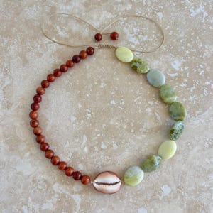 Op de afbeelding: Een ketting met een enkele kaurischelp in het midden, geflankeerd door een streng kleine, ronde, donkerbruine houten kralen aan de ene kant en een reeks grotere, gladde, ovaalvormige lichtgroene en gevlekte olijfgroene stenen kralen aan de andere.