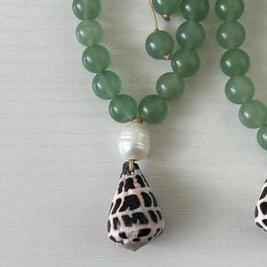 May include: A close-up of a necklace featuring green jade beads. The necklace has a white pearl and a shell pendant with black and brown markings. The necklace is on a white surface.