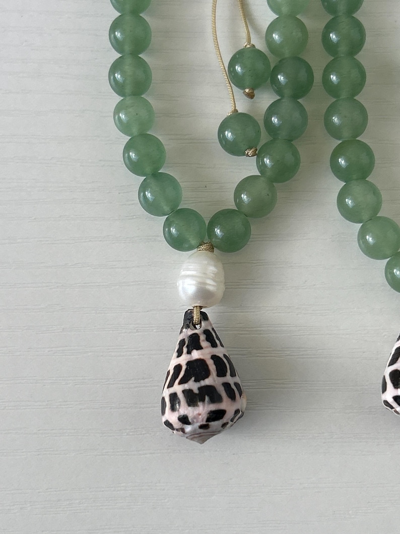 May include: A close-up of a necklace featuring green jade beads. The necklace has a white pearl and a shell pendant with black and brown markings. The necklace is on a white surface.