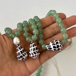 May include: A beaded necklace featuring light green round beads and three shell pendants. The pendants are white with black spotted patterns. One white pearl bead is also included in the design. The necklace is held in a hand.