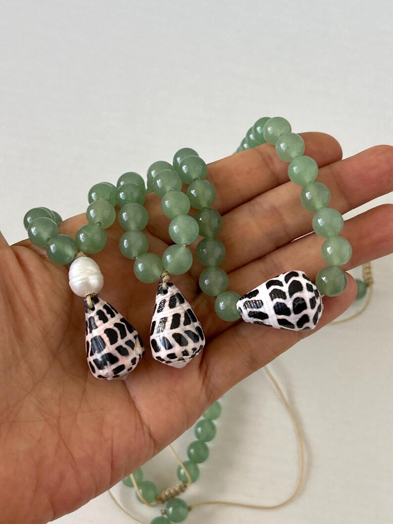 May include: A beaded necklace featuring light green round beads and three shell pendants. The pendants are white with black spotted patterns. One white pearl bead is also included in the design. The necklace is held in a hand.
