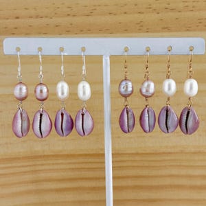 May include: Six pairs of earrings with a purple shell and a pearl dangle. The pearls are white and pink. The earrings are displayed on a white stand.