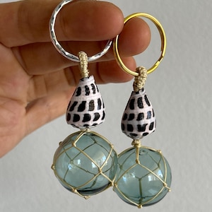 Könnte beinhalten: Zwei Schlüsselanhänger mit maritimem Thema. Jeder Schlüsselanhänger hat eine klare blaue Glaskugel, die in einem beigen Netz eingeschlossen ist, gekrönt von einer schwarz-weißen Muschel und einem silbernen oder goldenen Ring. Die Schlüsselanhänger werden gehalten.